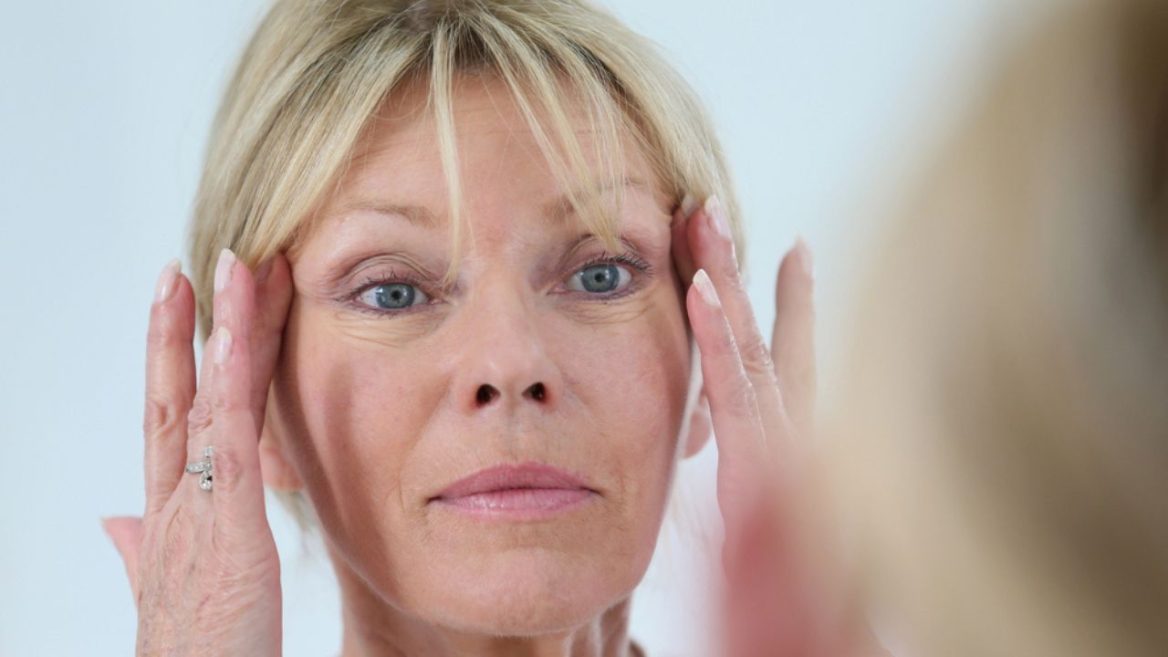 Femme mûre examinant sa peau dans un miroir, illustrant l'impact des traitements esthétiques tels que la mésothérapie et les injections d'acide hyaluronique sur le visage.